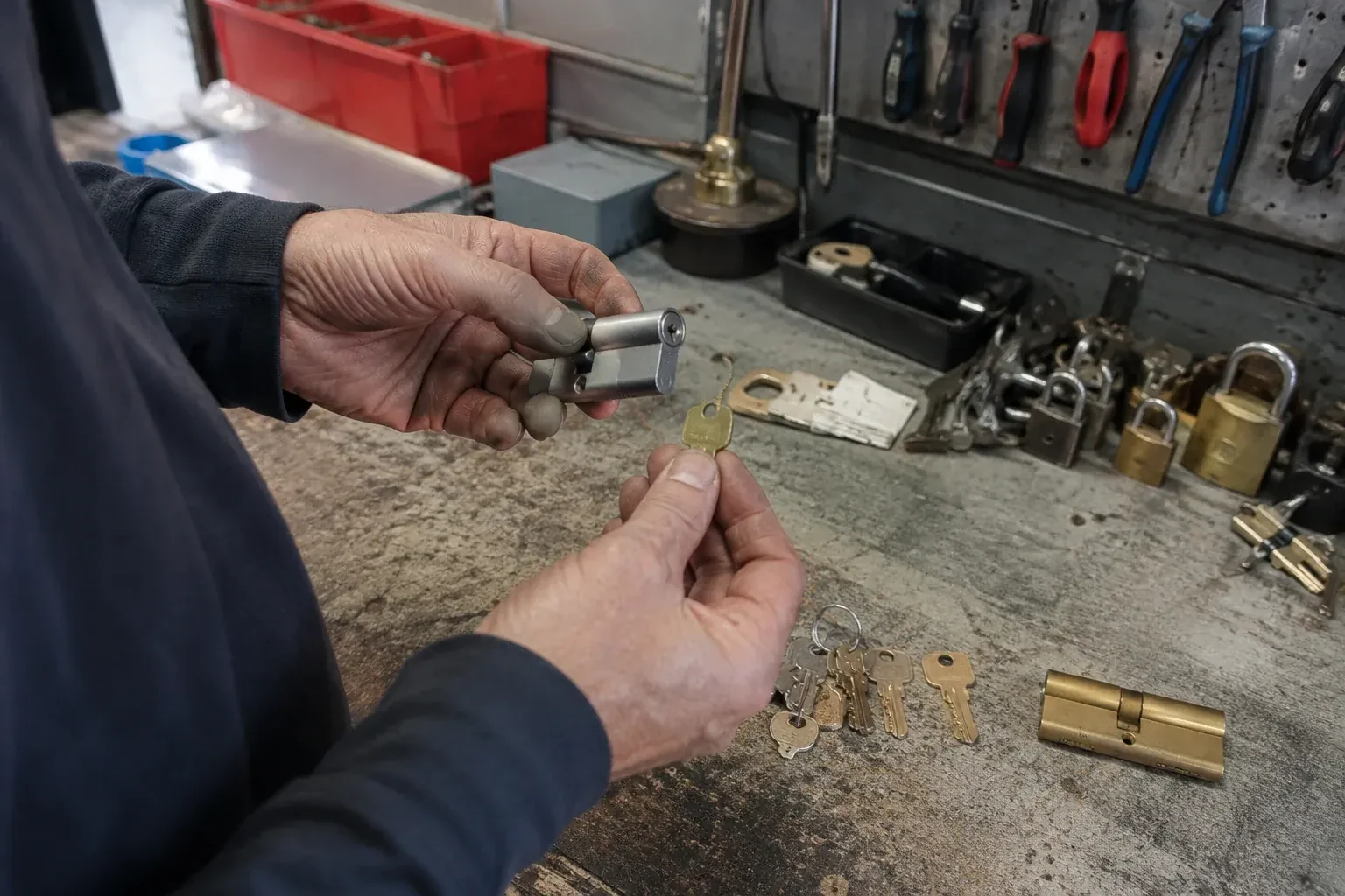 Hands holding a silver-colored lock cylinder and a brass-colored key above a workbench with padlocks, keys and tools. — Master Key System Design, Haldimand County